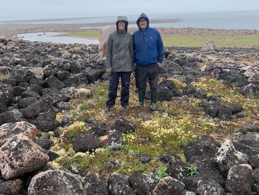 A woman and a man wearing hooded jackets stand in a mossy area surrounded by a ring of rocks.