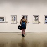 Woman looking at a piece of art in a gallery. Four other artworks are visible.