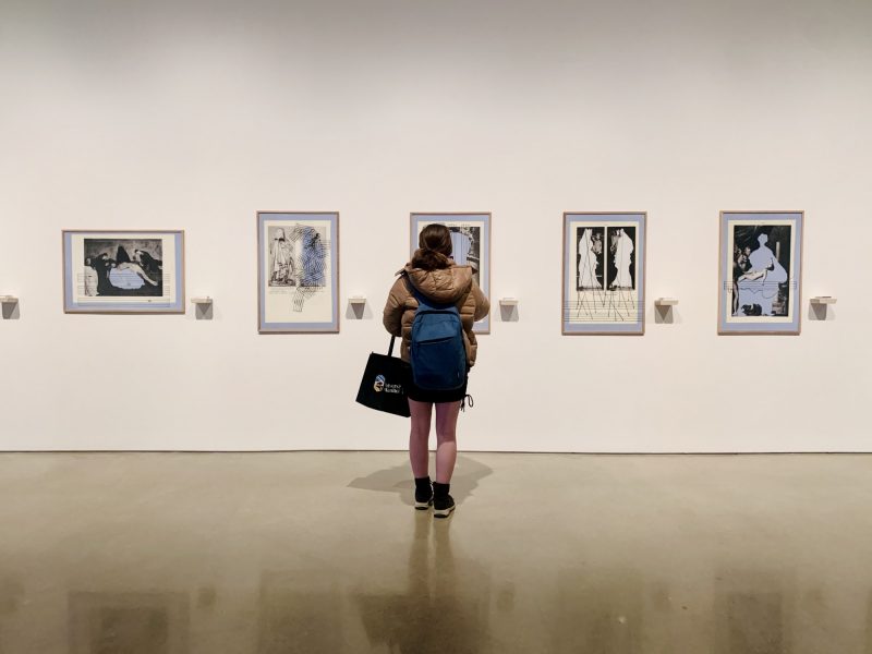 Woman looking at a piece of art in a gallery. Four other artworks are visible.