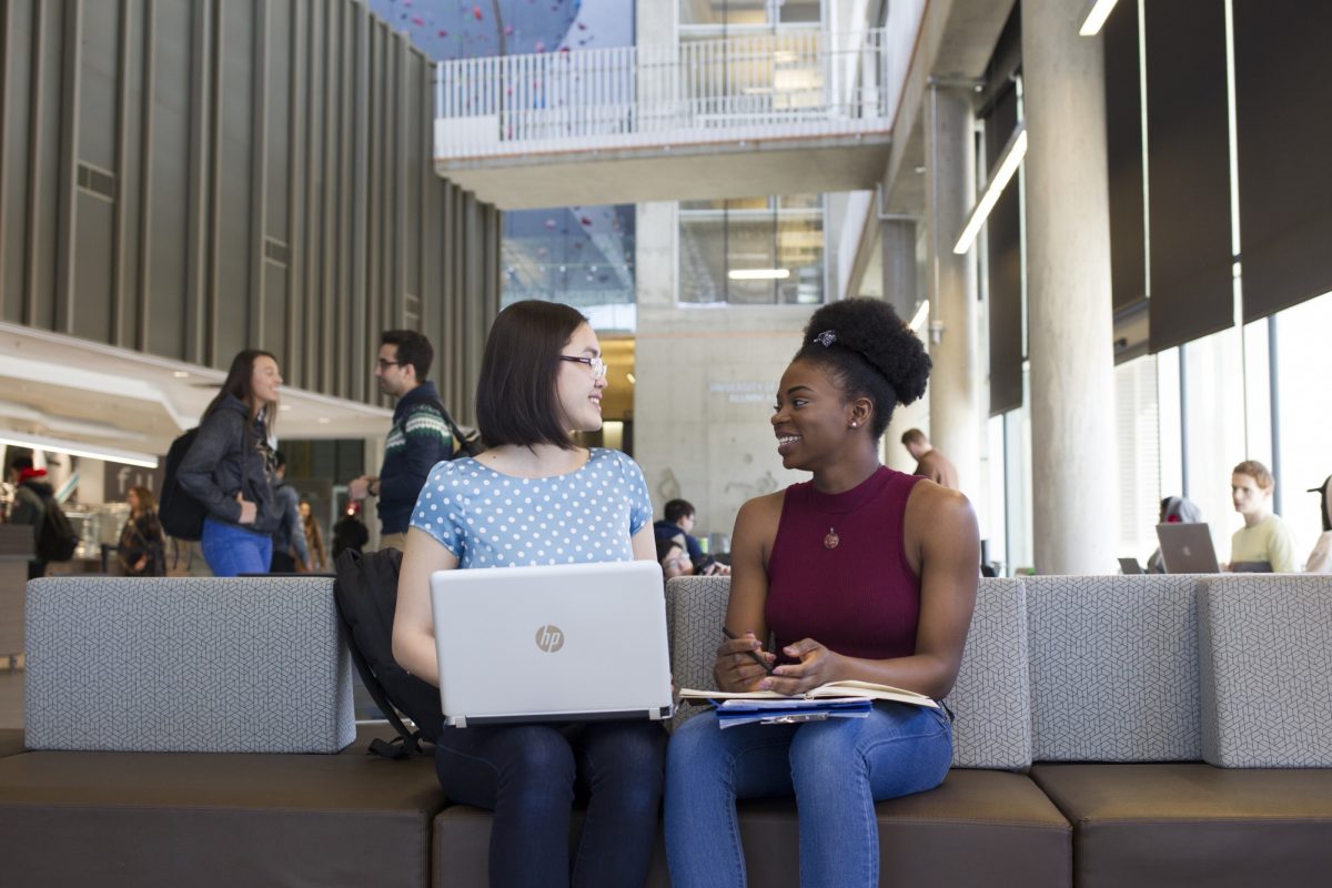 Two smiling people sit on a bench chatting with each other. One has a laptop in her lap while the other has a notebook.