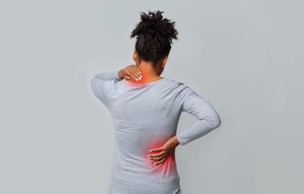 A standing woman is seen from behind, with one hand on her neck and one on her lower back, with both areas illuminated in red to show pain.