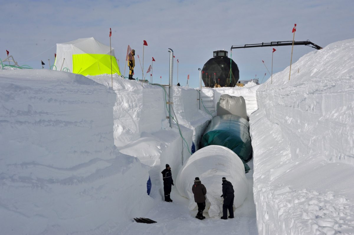 A large snow trench being built. Three people are visible in the trench, along with three giant balloons. A crane and bright yellow tent sit above the trench.