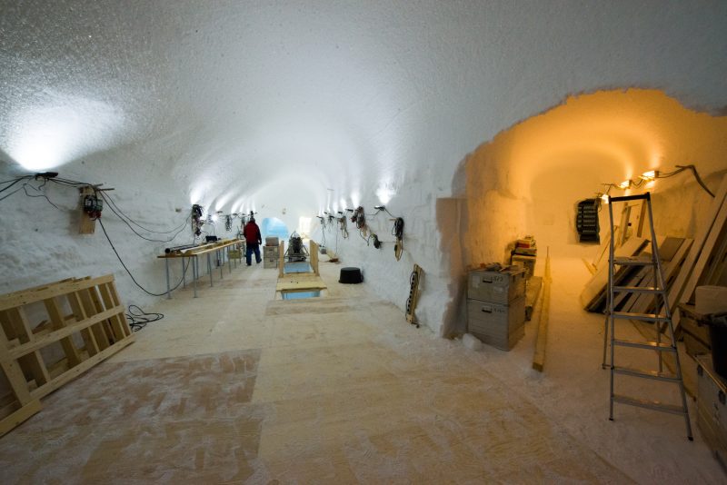 A hallway-sized tunnel in the snow, with plywood floors, and lights installed near the ceiling. A person walks near the end of the tunnel.