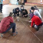 Students holding a web-like string on wood floor.