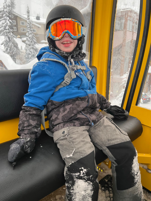 A boy in snow gear sits on a bench, smiling.