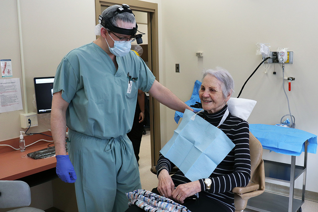 Catherine sits in a dental chair and wears a bib. Dr. McCoy stands next to her and wears scrubs, gloves and a face mask.