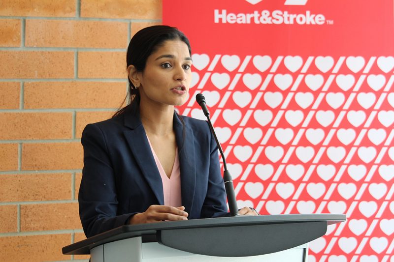 Dr. Nishita Singh speaks into a microphone at a lectern. The Heart & Stroke logo and the words "Heart & Stroke" are on a banner behind her.