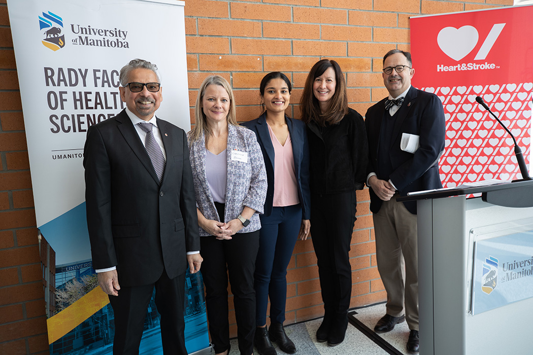 The partners pose for a photo. A banner behind them reads "Heart & Stroke" with its logo. Another banner behind them reads "University of Manitoba. Rady Faculty of Health Sciences" with UM logo.