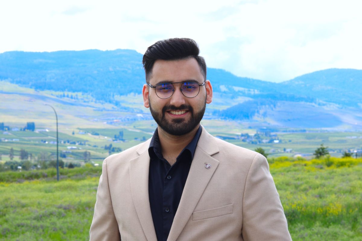 Man in beige suit and black shirt with smiling in front of a green and blue hillscape.