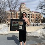 Darby Goodall, Science alumni, standing in front of the Administration Building in a black dress holding photos of her running accomplishments