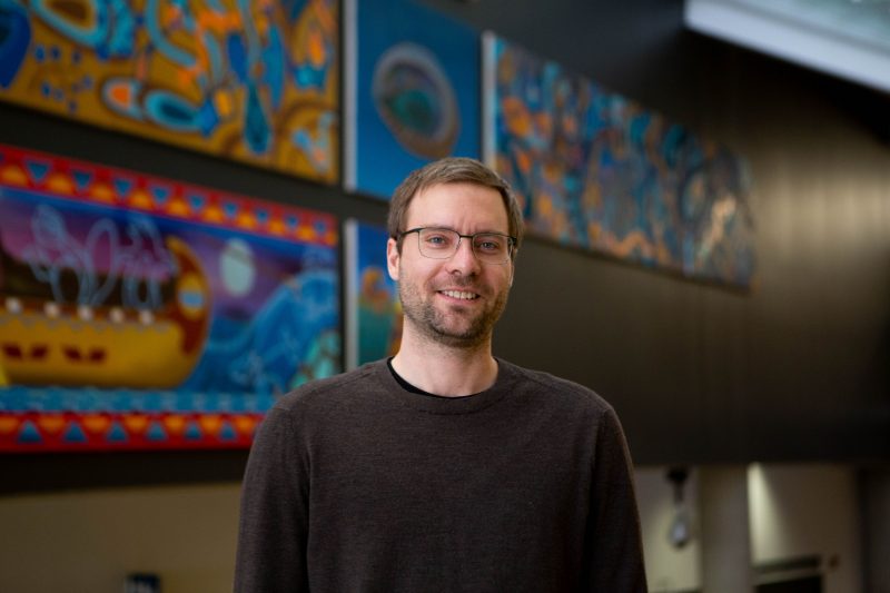 Man with short brown hair, close shaved brown beard wearing glasses, wearing a brown sweater.