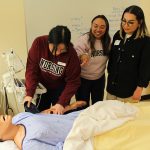 Three female nursing students practice skills in a simulated hospital room with a manikin.