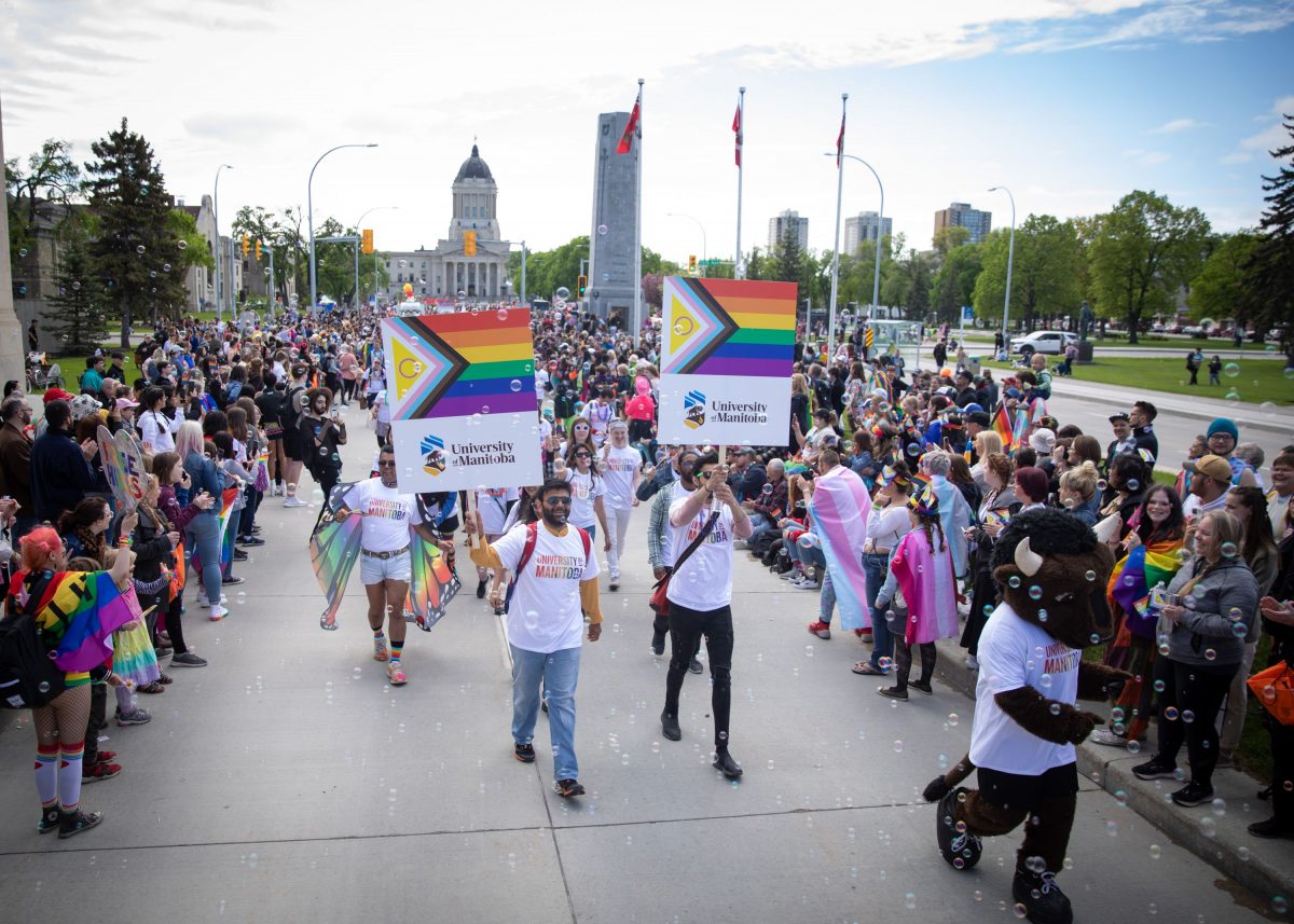 crowd of people walking in pride parade on sunny day, holding university of manitoba signs with pride colours