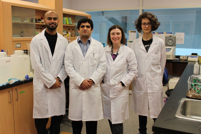 The professor and his grad students pose for a photo in his lab. They are wearing lab coats. 