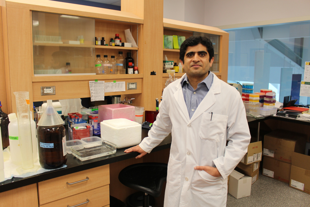 Portrait of Dr. Tanveer Sharif in his lab. He is wearing a lab coat. There is scientific equipment and bottles on the counter and on shelves behind him.