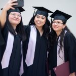 Three students wearing graduation caps and gowns. One student holds a smartphone and takes a selfie with the other two students.
