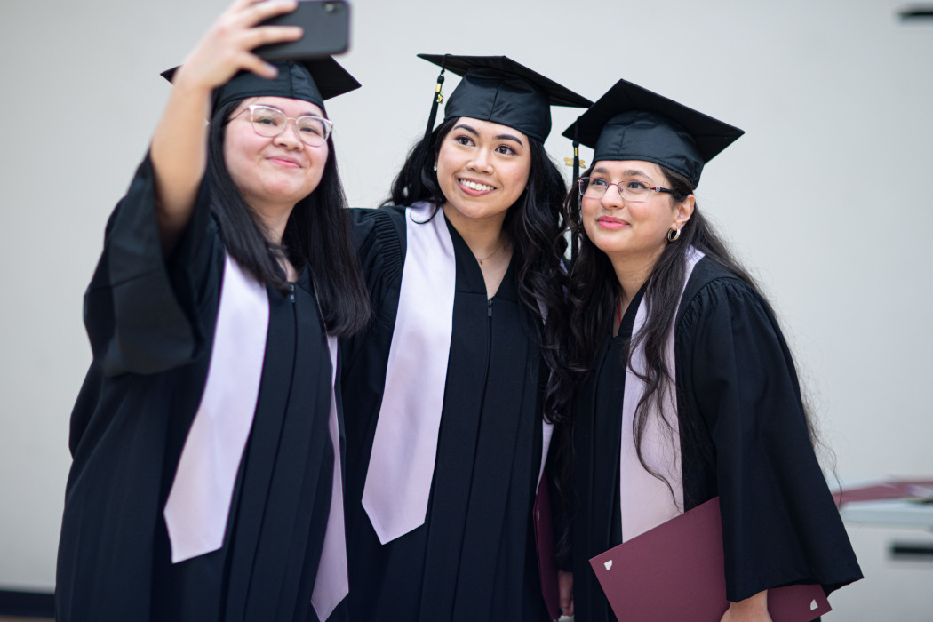 Three students wearing graduation caps and gowns. One student holds a smartphone and takes a selfie with the other two students.