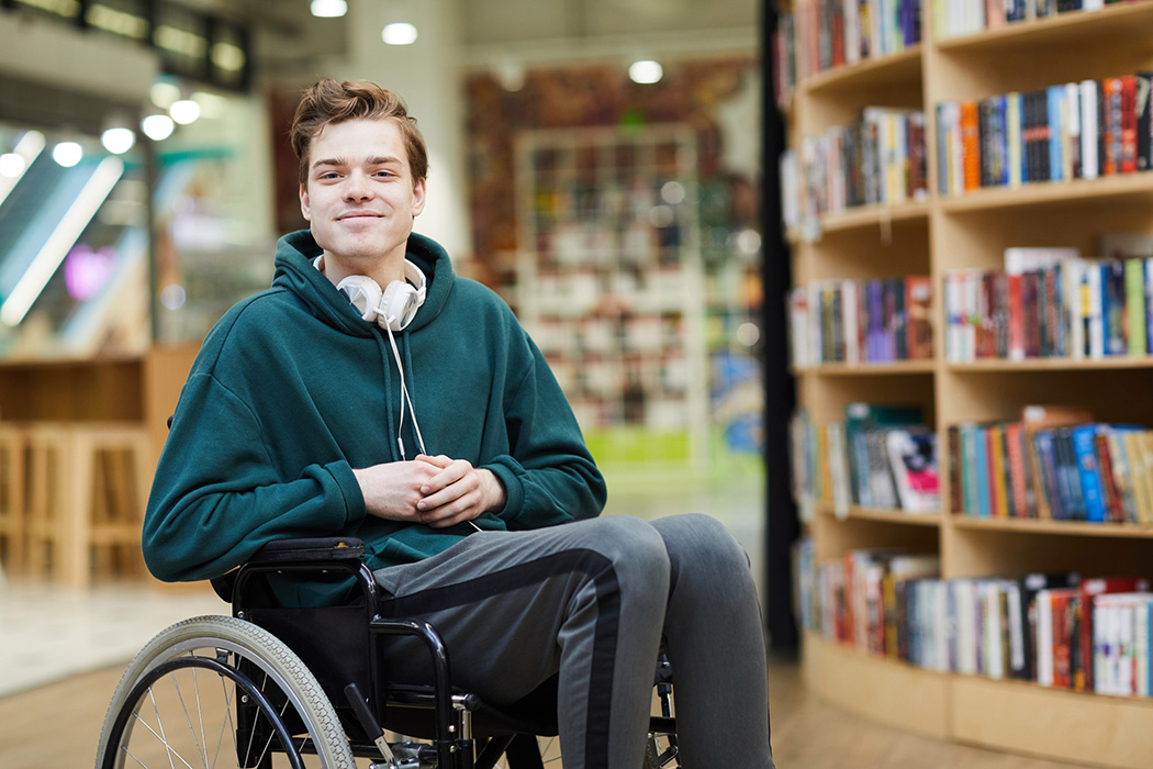 Content handsome young disabled student with headphones on neck siting in wheelchair and looking at camera in modern library or bookstore.