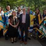Eight of ten of the 2023 Honouring Our Indigenous Campus Community honourees stand with UM Vice-President (Indigenous) Dr. Catherine Cook and UM President Dr. Michael Benarroch. The honourees are wrapped in Star Blankets and they are all standing outside in front of some trees.