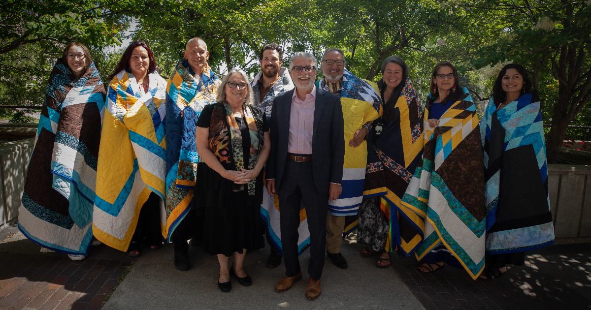 Eight of ten of the 2023 Honouring Our Indigenous Campus Community honourees stand with UM Vice-President (Indigenous) Dr. Catherine Cook and UM President Dr. Michael Benarroch. The honourees are wrapped in Star Blankets and they are all standing outside in front of some trees.