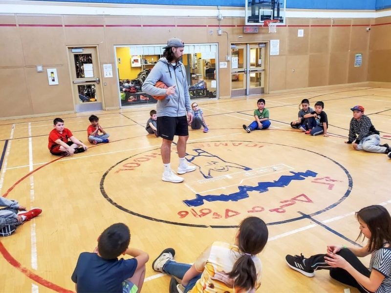 Keegan Slijker standing in the middle of a circle of sitting children, teaching basketball.