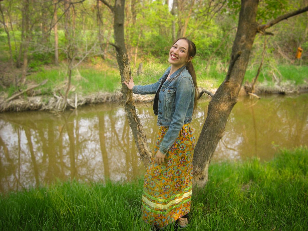 Female wearing a ribbon skirt and jean jacket standing in front of a river.