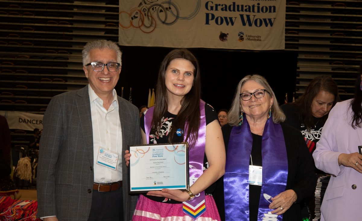 A UM graduate stands between the UM President and Vice-President (Indigenous), holding her diploma, at the graduation pow wow.