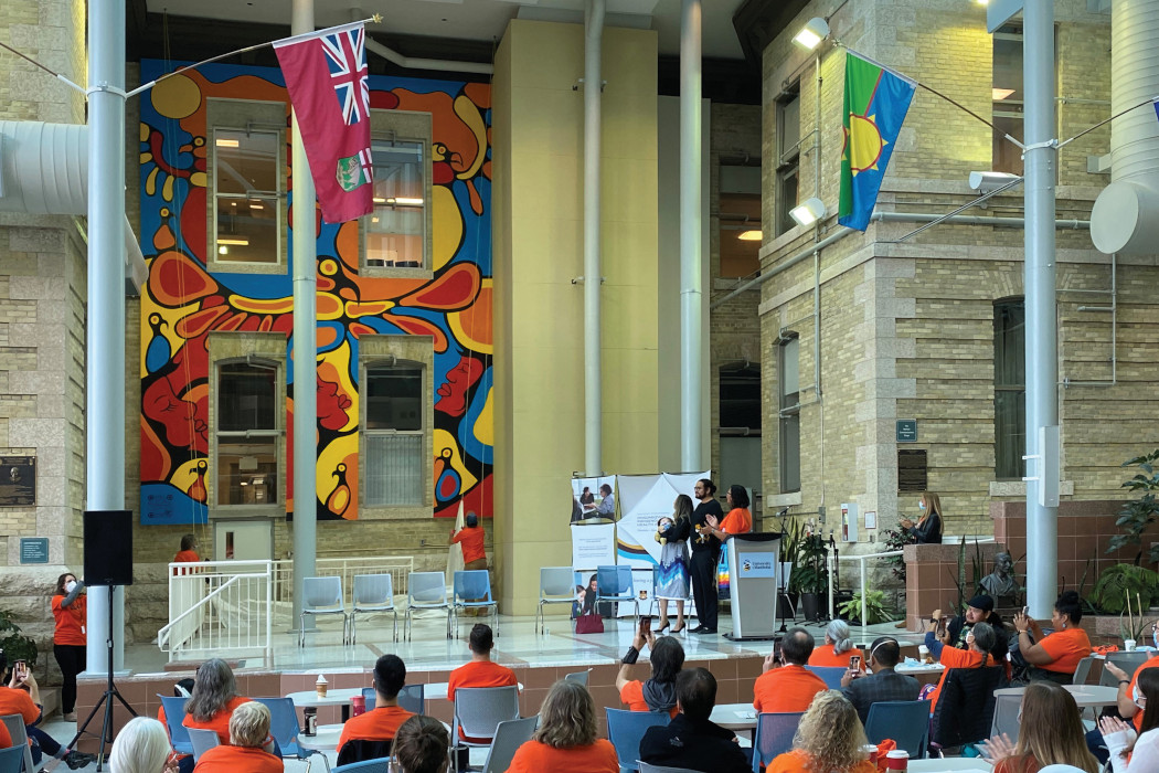 Mural featuring Indigenous symbols is unveiled. Blake Angeconeb stands near a lectern on the stage. People in the crowd are wearing orange shirts and some are taking photos of the mural.