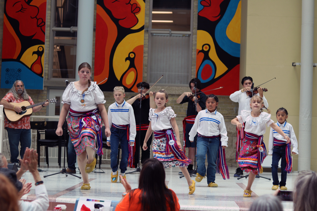 Six youths dance while three people play fiddles and one man plays guitar.