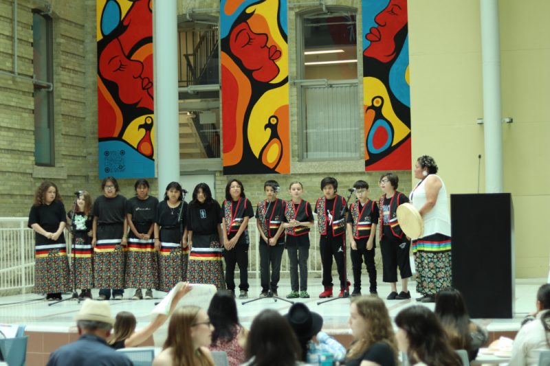 12 kids sing on stage. A woman stands next to them holding a drum. 