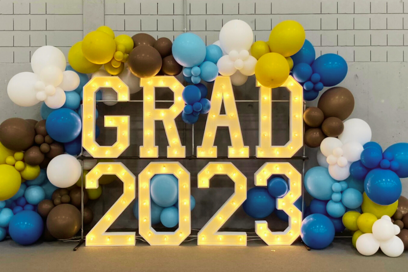 marquee letters spelling "grad 2023" surrounded by white, brown, gold and blue balloons