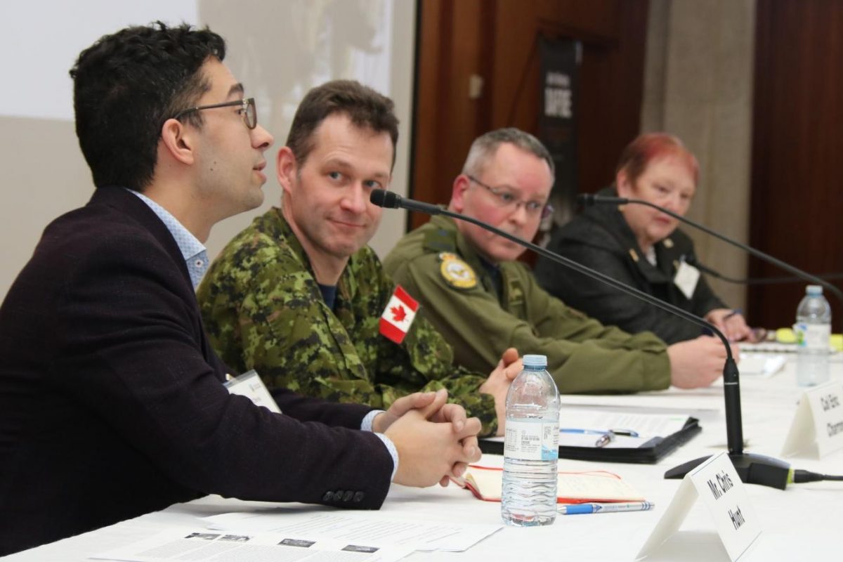 Four people sitting at table with microphones as they are on a speaker's panel. Two men are dressed in military uniforms.