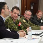 Four people sitting at table with microphones as they are on a speaker's panel. Two men are dressed in military uniforms.