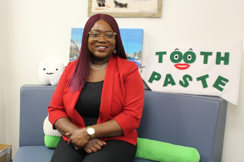 Dr. Olubukola-Olatosi sits on a small couch. Behind her is a sign that reads "tooth paste." The two "o's" in tooth form eyes and there are eyebrows above them and a mouth below. A smiling plush tooth with one tooth is also behind her.