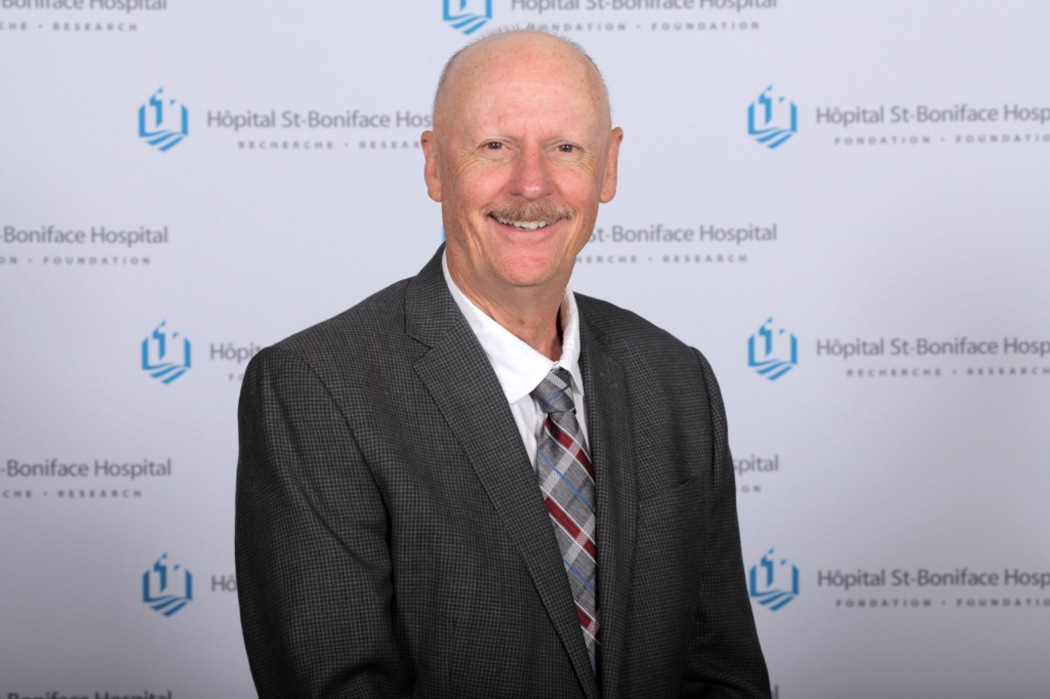 Portrait of Dr. Grant Pierce. In the background is the St. Boniface Hospital logo and it reads "Hopital St-Boniface Hospital, Recherche, Research," and "Foundation."