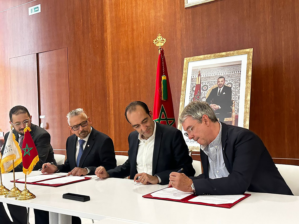 four men in suits, seated at a table with one man signing a document while the others watch