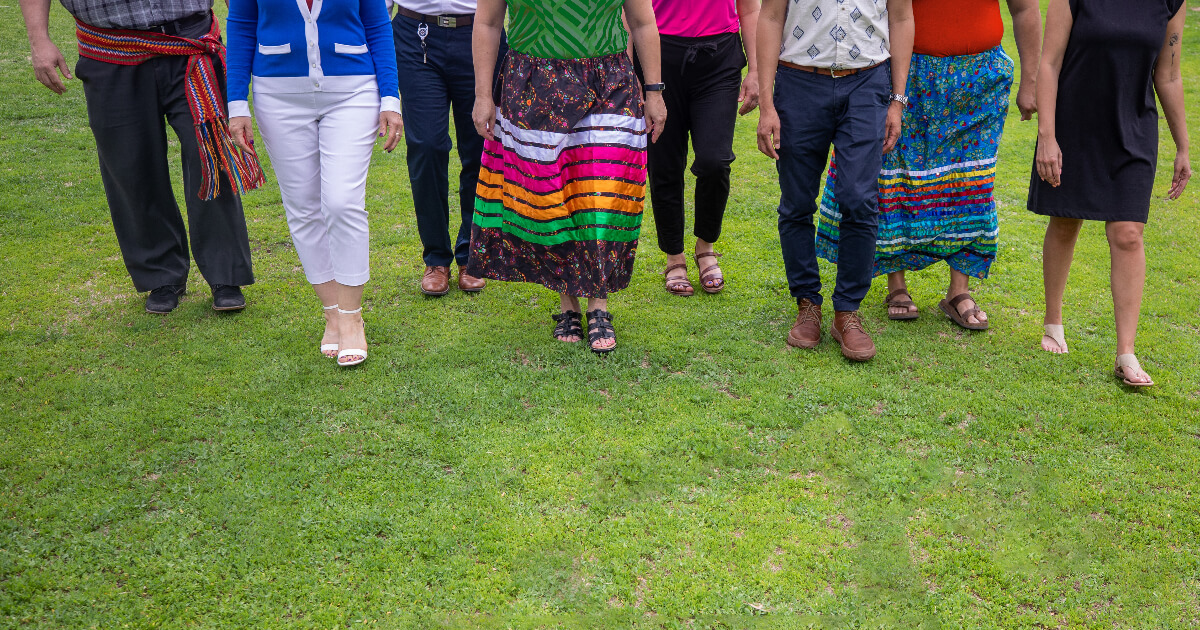 Seven people walking together, shown from the waist down, with two wearing ribbon skirts and one in a Métis sash.