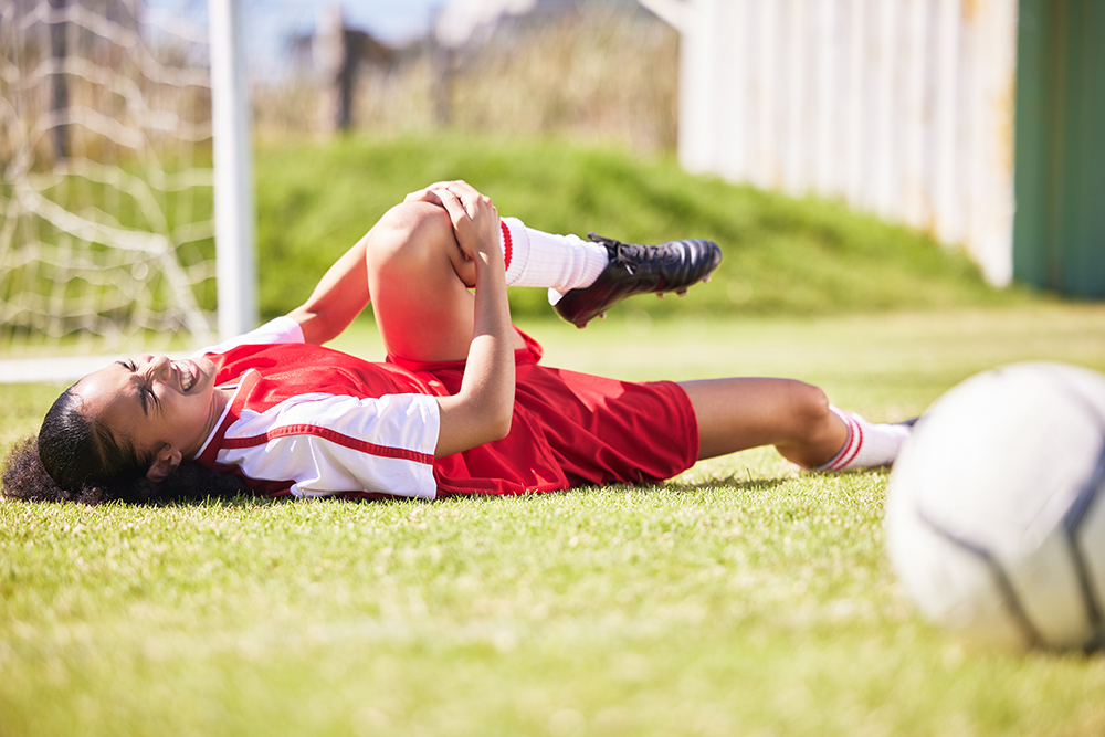 A female soccer player lying on a field holding her leg on the ground in agony.