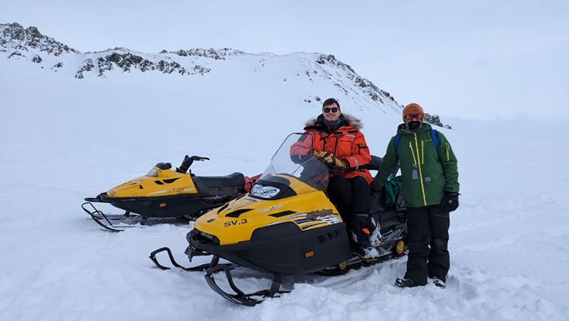 Two researchers standing next to their snowmobiles in snow.