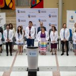 10 medical students stand on stage wearing white coats. Faculty members and special guests stand behind the students.