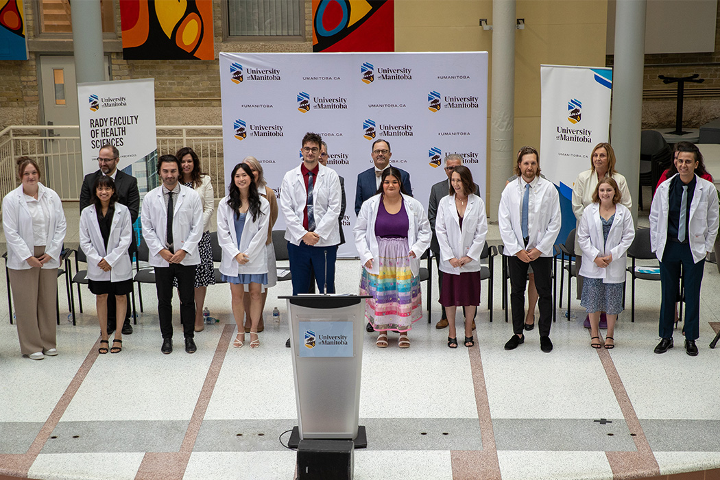 10 medical students stand on stage wearing white coats. Faculty members and special guests stand behind the students.