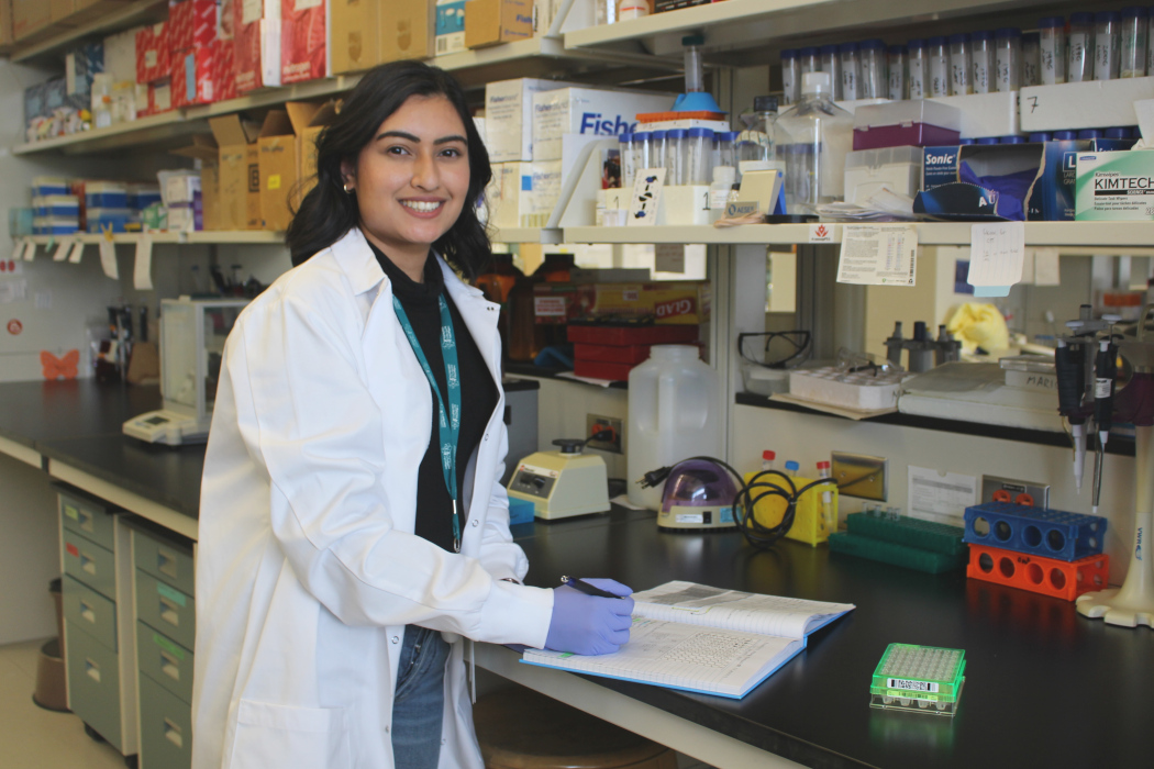 Portrait of Khushali Trivedi in a lab. She is wearing a lab coat. She is holding a pen and a note book is on the counter.