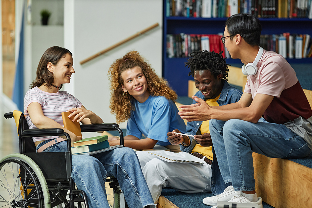 A diverse group of young people chatting in college library including female student with disability.