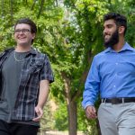 2023 Schulich Leaders Cody McDonald and Simar Ubhi walk across campus. They are smiling as they talk to each other and there are green trees in the background. Cody wears glasses and dark clothing under a plaid shirt. Simar wears a blue dress shirt with khakis.
