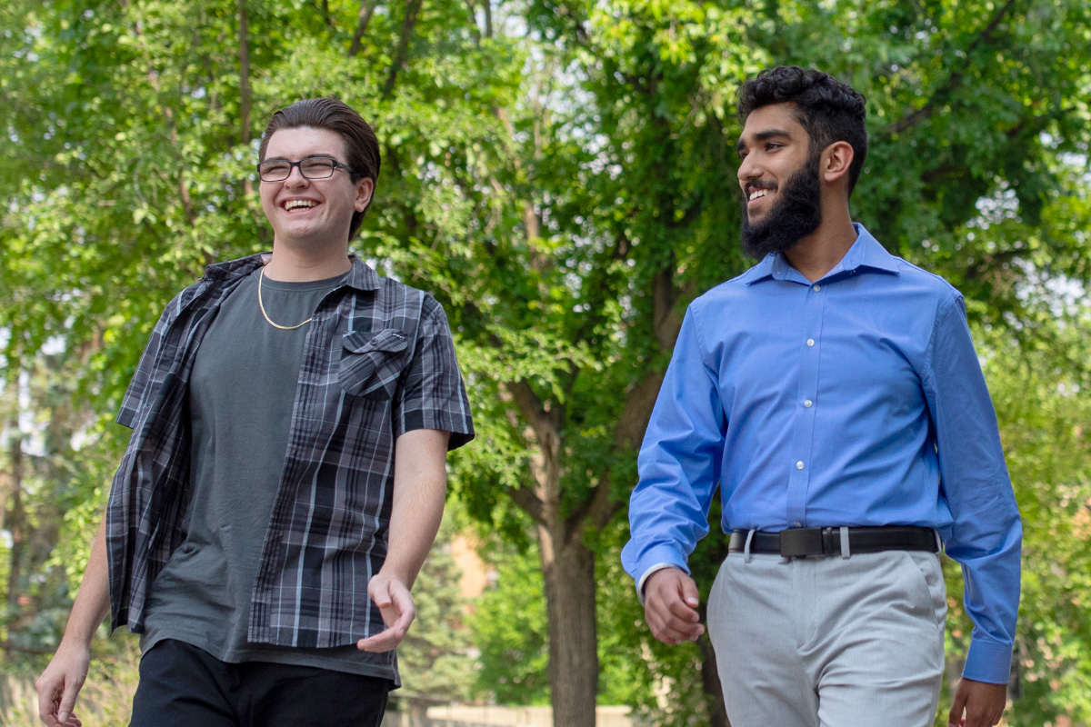 2023 Schulich Leaders Cody McDonald and Simar Ubhi walk across campus. They are smiling as they talk to each other and there are green trees in the background. Cody wears glasses and dark clothing under a plaid shirt. Simar wears a blue dress shirt with khakis.