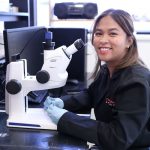 Student Deanne Nixie Miao smiles while sitting at a microscope in a lab.