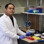 Erwin Taguiam, wearing a white lab coat, holds a small test tube in a lab.