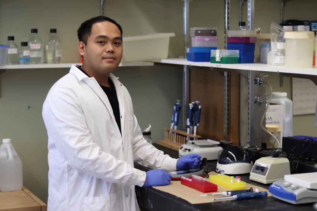 Erwin Taguiam, wearing a white lab coat, holds a small test tube in a lab.