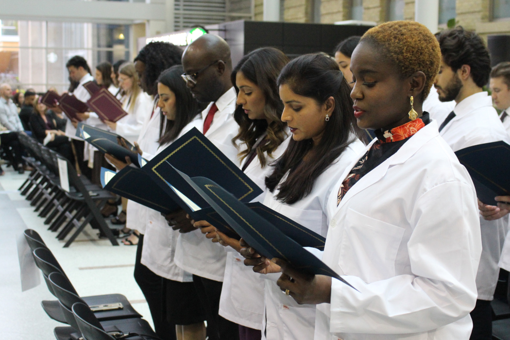 Rows of students in white coats are standing. They are each holding an open folder, and reading the Community Code off a piece of paper.