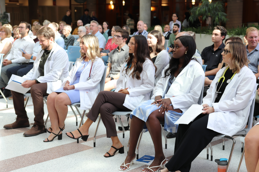 Five MPAS students wearing white coats and stethoscopes are sitting in chairs. A crowd of people is sitting behind them.